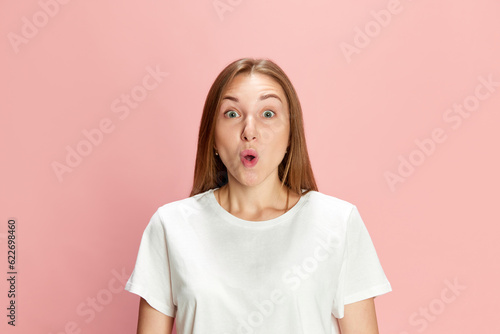 Portrait of beautiful young woman in casual white t-shirt posing with shocked face against pink studio background. Cheerful surprise