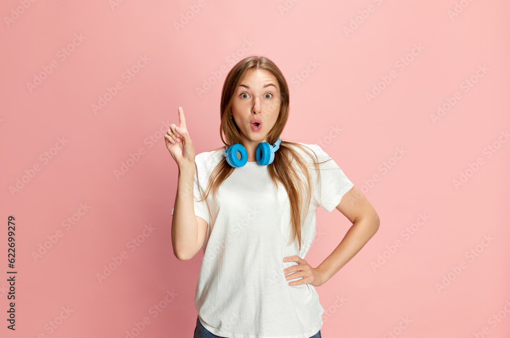 Portrait of young beautiful girl in casual clothes and headphones raising finger up against pink studio background. Ideas Portrait of young beautiful girl in casual clothes and headphones raising finger up against pink studio background. Ideas