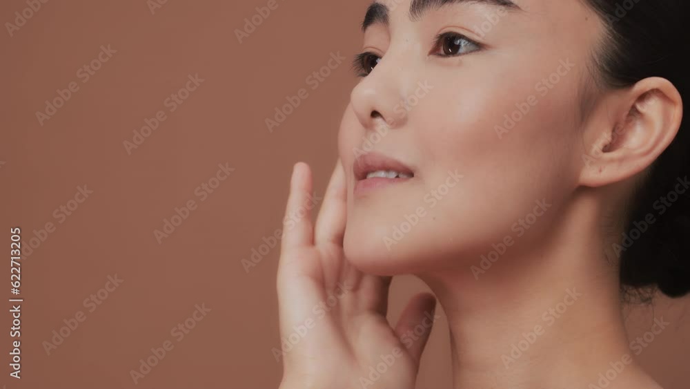 Close-up side view of a beauty asian young touches her face, apply the cream, smiling on beige background. Beautiful female model with perfect clean fresh skin. Skin care treatment concept Close-up side view of a beauty asian young touches her face, apply the cream, smiling on beige background. Beautiful female model with perfect clean fresh skin. Skin care treatment concept