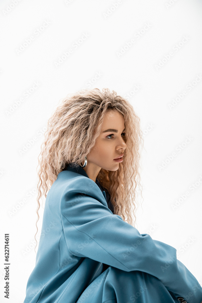profile portrait of mesmerizing woman with wavy ash blonde hair wearing blue stylish blazer and looking away on grey background, charming and sensual female model, modern fashion