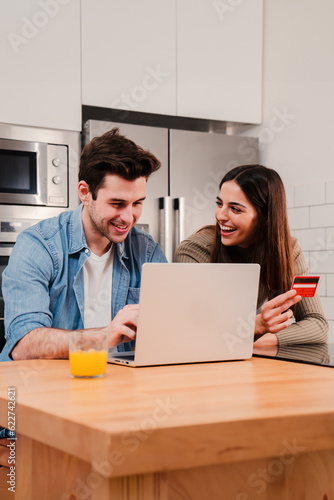 Vertical portrait of a couple smiling using a laptop and a credit card to buy on internet. Young husband and happy wife paying a online shopping purchase. Boyfriend and girlfriend ordering a delivery