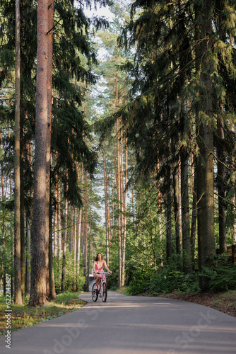 young active woman in pink dress rides a bicycle in park among the trees. Women fun weekend sports and recreation. Walking along bike path in national park, freedom and speed. riding