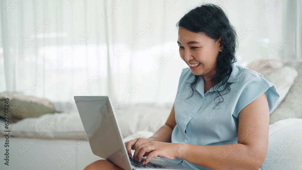 Happy adult asian women with prosthetic leg typing on laptop for working online or chatting sitting on comfortable sofa at bedroom. Leg prosthetic equipment, Amputee concept Happy adult asian women with prosthetic leg typing on laptop for working online or chatting sitting on comfortable sofa at bedroom. Leg prosthetic equipment, Amputee concept
