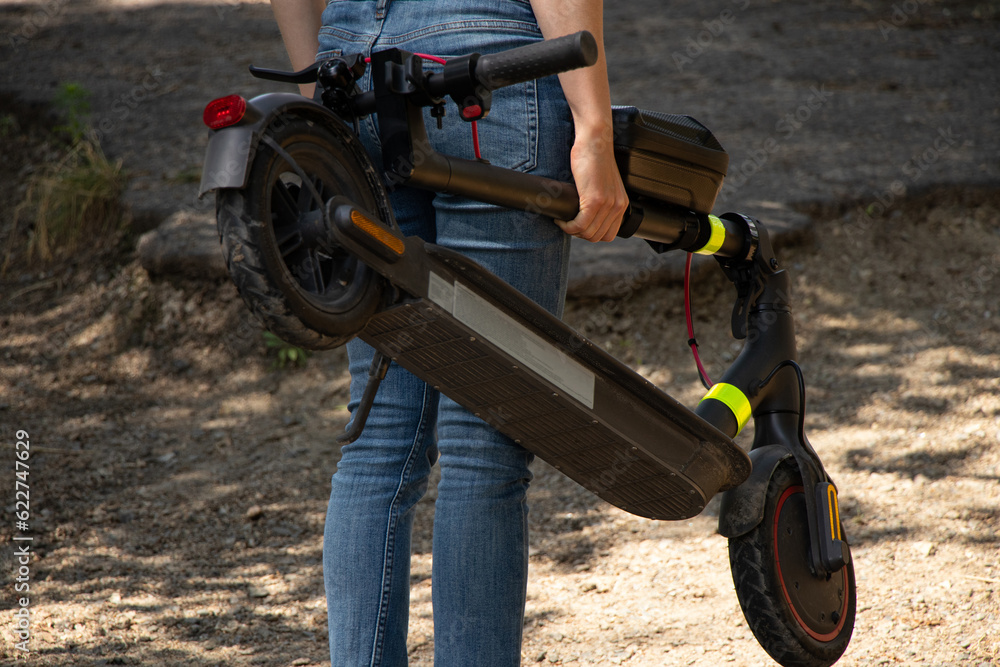 Obraz premium A girl holds an electric scooter in her hands in parks in Ukraine in the summer, urban transport