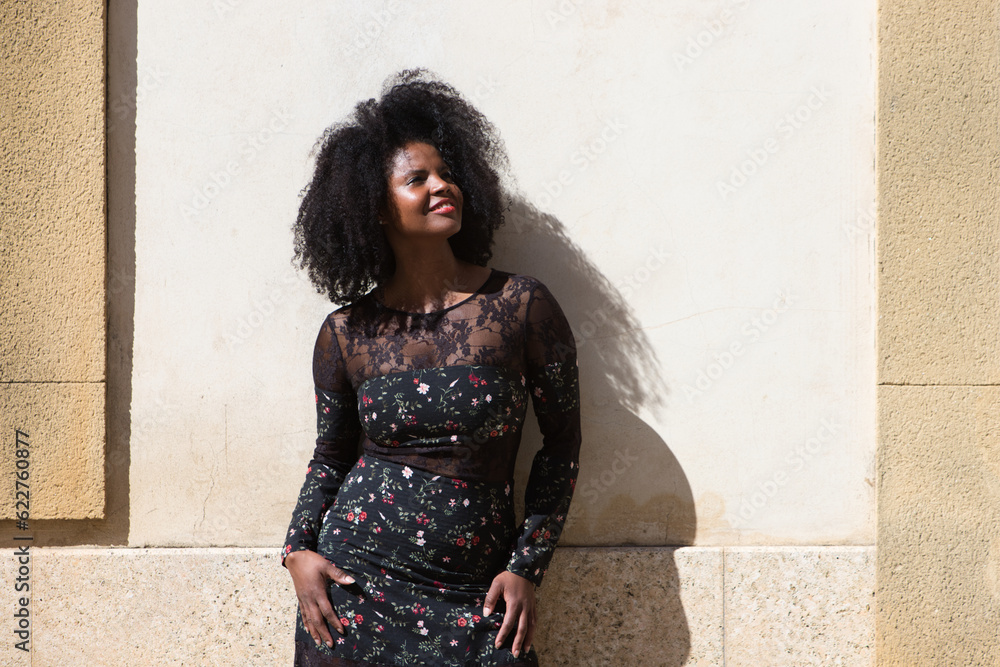 Portrait of black woman, young and beautiful with afro hair and black dress with flowers is very happy and smiling. In the background a light brown wall. Concept different expressions. Portrait of black woman, young and beautiful with afro hair and black dress with flowers is very happy and smiling. In the background a light brown wall. Concept different expressions.