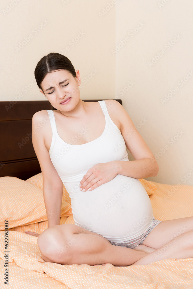 Young pretty pregnant woman, in pain, sitting on bed in bedroom, preparing to give a birth Young pretty pregnant woman, in pain, sitting on bed in bedroom, preparing to give a birth