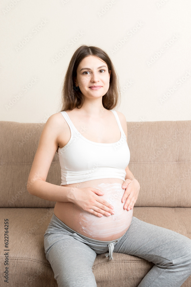 a pregnant girl sits at home on the bed and smears an anti-stretch mark cream on her stomach. Pregnancy, motherhood, preparation and expectation concept a pregnant girl sits at home on the bed and smears an anti-stretch mark cream on her stomach. Pregnancy, motherhood, preparation and expectation concept
