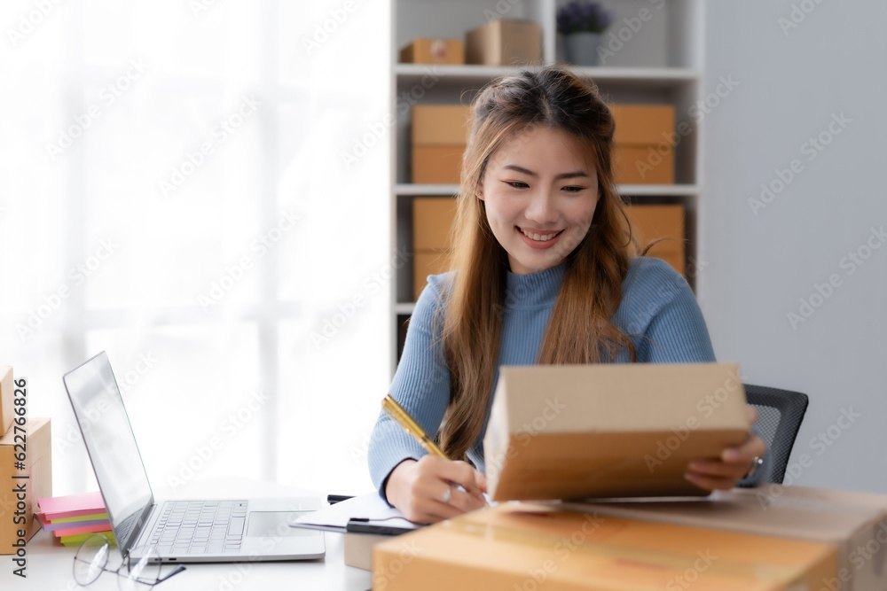 Smiling young Asian business owner woman prepare parcel box and check online orders of product for deliver to customer on laptop computer at home. Shopping Online concept. Startup small business SME. Smiling young Asian business owner woman prepare parcel box and check online orders of product for deliver to customer on laptop computer at home. Shopping Online concept. Startup small business SME.