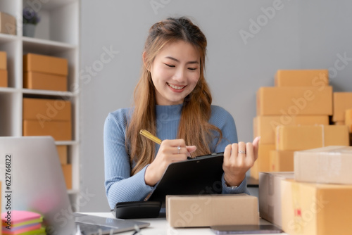 Smiling young Asian business owner woman prepare parcel box and check online orders of product for deliver to customer on laptop computer at home. Shopping Online concept. Startup small business SME.