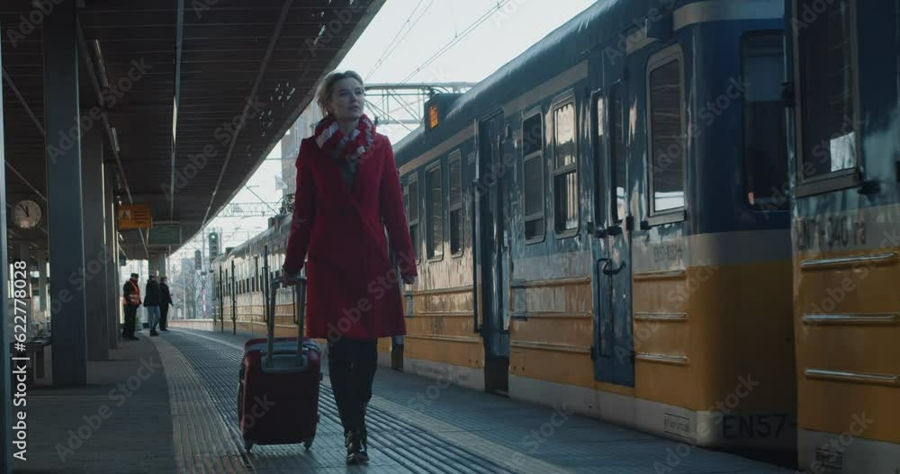 Woman in a Red Coat Traveler with Trolley Bag Walking Along the Station Platform Along the Departing Modern Train. She's About to go on a Train Journey Woman in a Red Coat Traveler with Trolley Bag Walking Along the Station Platform Along the Departing Modern Train. She's About to go on a Train Journey