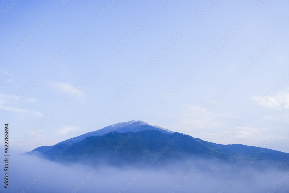 Beautiful view of Mount Merbabu mountainside in the morning with vegetable garden and village settlements or housing in Selo Boyolali, Central Java, Indonesia. Blue sky, fog, mist, clouds backgrounds