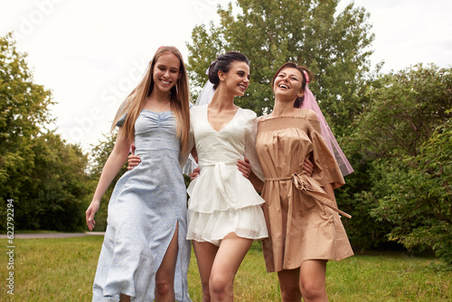 Happy adorable bride in a white dress with white veil and two bridesmaids in blue and beige dress with pink veils walking in a summer park hugging each other. Cheerful bachelorette party. © Georgii
