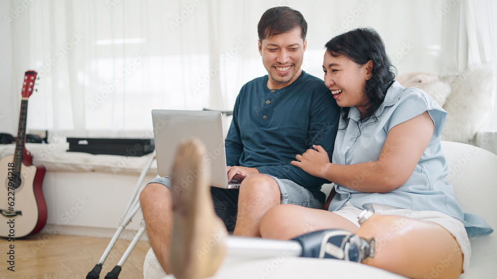 Happy asian woman in leg prosthesis and her husband sitting on comfortable sofa while typing on laptop for working online or chatting on social media at home. Leg prosthetic equipment Happy asian woman in leg prosthesis and her husband sitting on comfortable sofa while typing on laptop for working online or chatting on social media at home. Leg prosthetic equipment