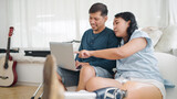 Happy asian woman in leg prosthesis and her husband sitting on comfortable sofa while typing on laptop for working online or chatting on social media at home. Leg prosthetic equipment