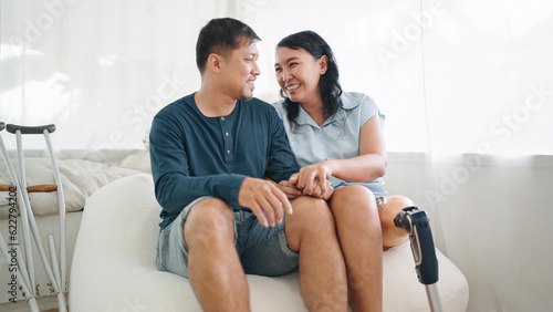 Happy asian woman in leg prosthesis and her husband sitting on comfortable sofa spending time together at home. Happy asian couple  Leg prosthetic equipment