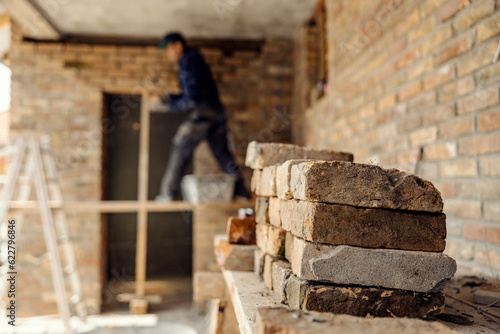 Small pile of bricks on a construction site with a worker in a background building a house.