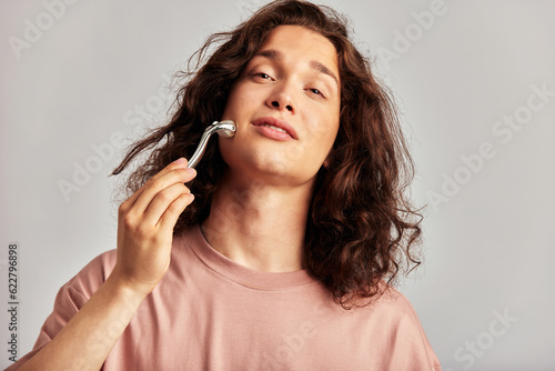 Young handsome long haired guy using de-puffing face roller to keep his skin soft and smooth. Caucasian man with brown hair maintaining his natural beauty with skincare routine. Men s self-care.