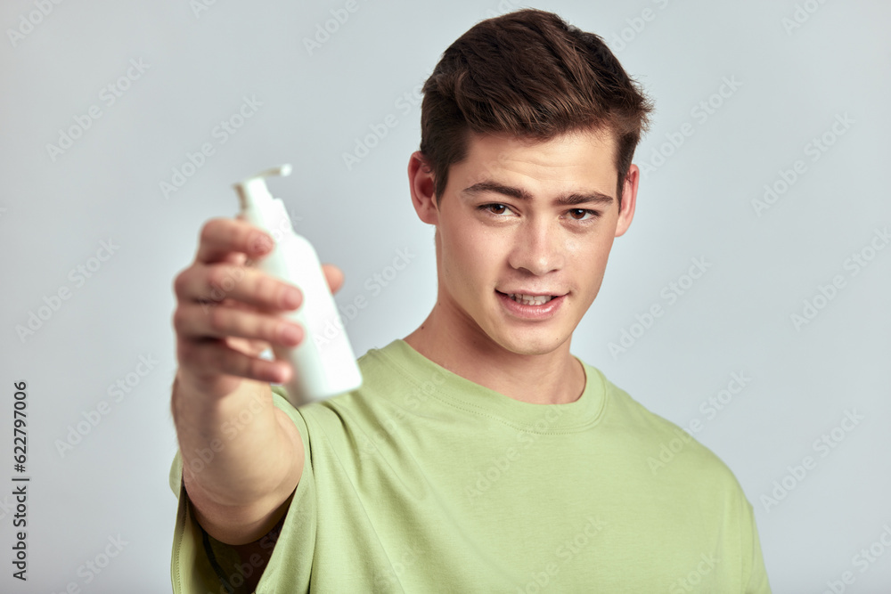 Young handsome smiling brown haired guy holding a bottle of facial wash or beauty cream in his hand. Caucasian millennial man uses hydrating skin products in his skincare routine to keep healthy look. Young handsome smiling brown haired guy holding a bottle of facial wash or beauty cream in his hand. Caucasian millennial man uses hydrating skin products in his skincare routine to keep healthy look.