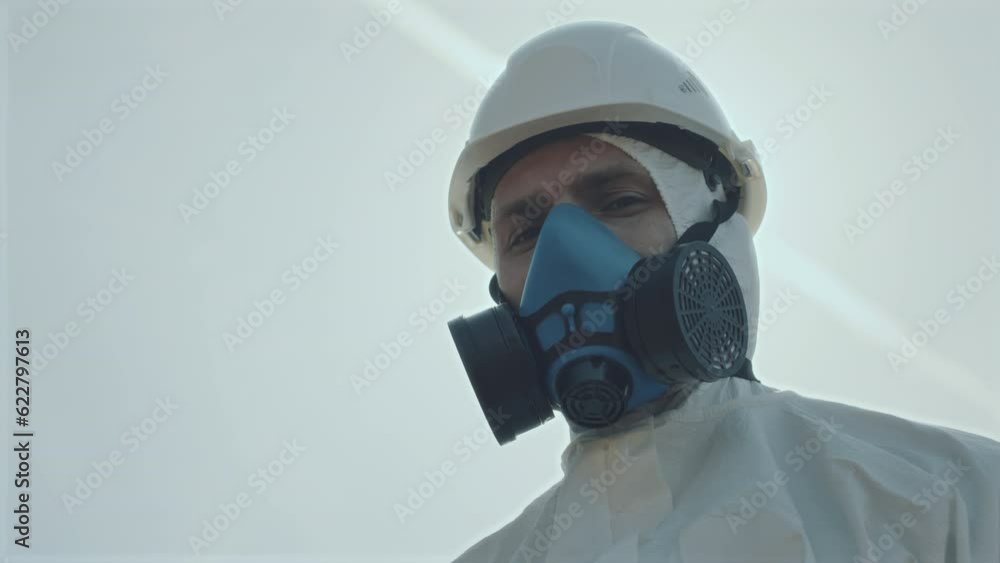Low angle portrait shot of male ecologist in protective coverall and respiratory mask standing outdoors and posing for camera Low angle portrait shot of male ecologist in protective coverall and respiratory mask standing outdoors and posing for camera