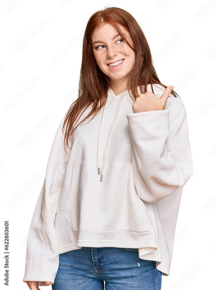 Young read head woman wearing casual sweatshirt smiling with happy face looking and pointing to the side with thumb up. Young read head woman wearing casual sweatshirt smiling with happy face looking and pointing to the side with thumb up.