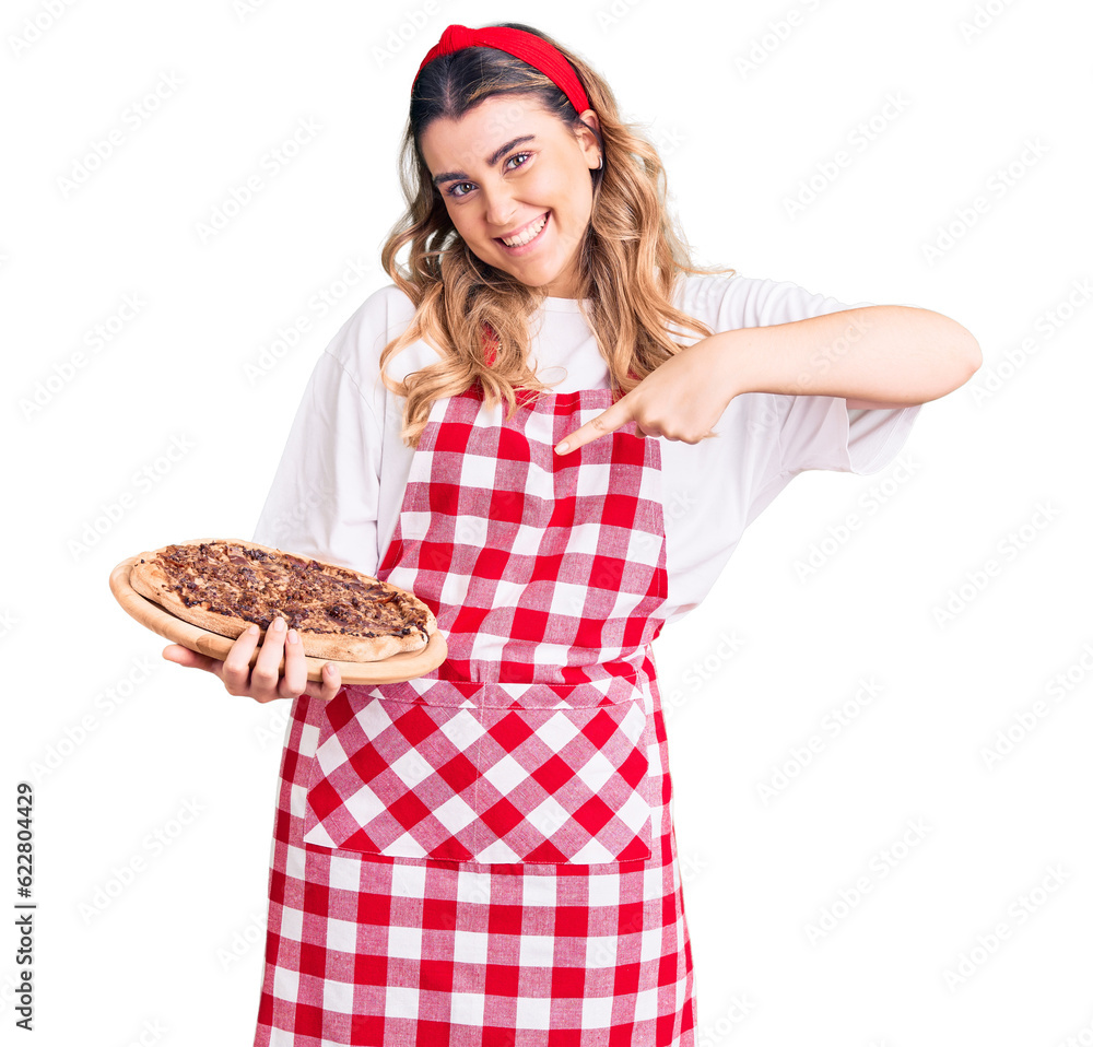 Young caucasian woman wearing apron holding pizza smiling happy pointing with hand and finger Young caucasian woman wearing apron holding pizza smiling happy pointing with hand and finger