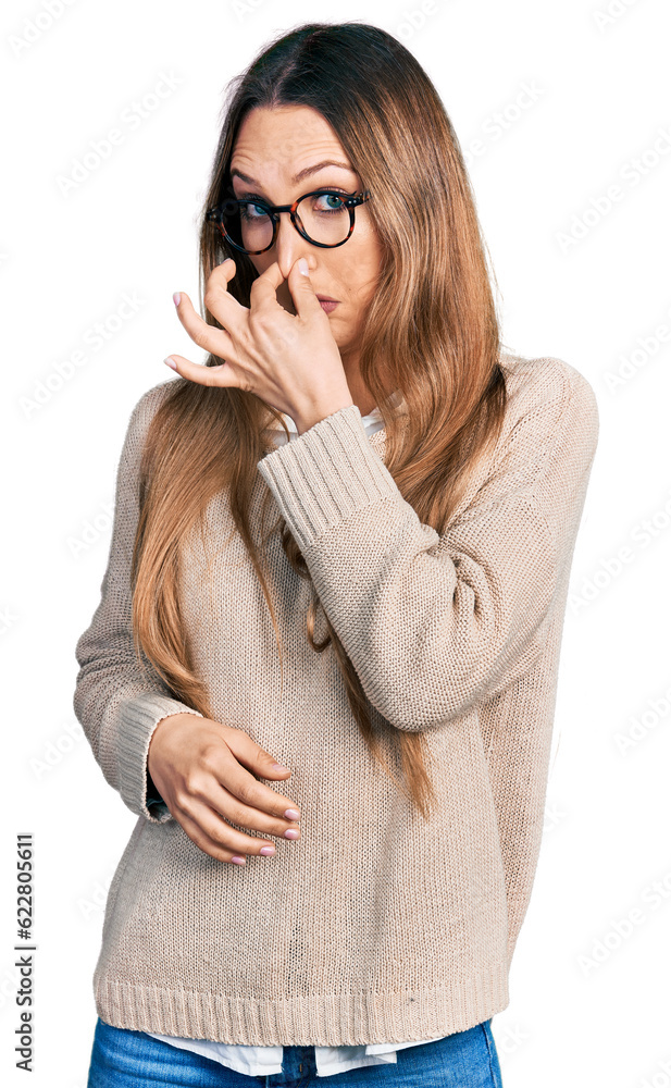 Beautiful caucasian woman wearing casual sweater and glasses smelling something stinky and disgusting, intolerable smell, holding breath with fingers on nose. bad smell
