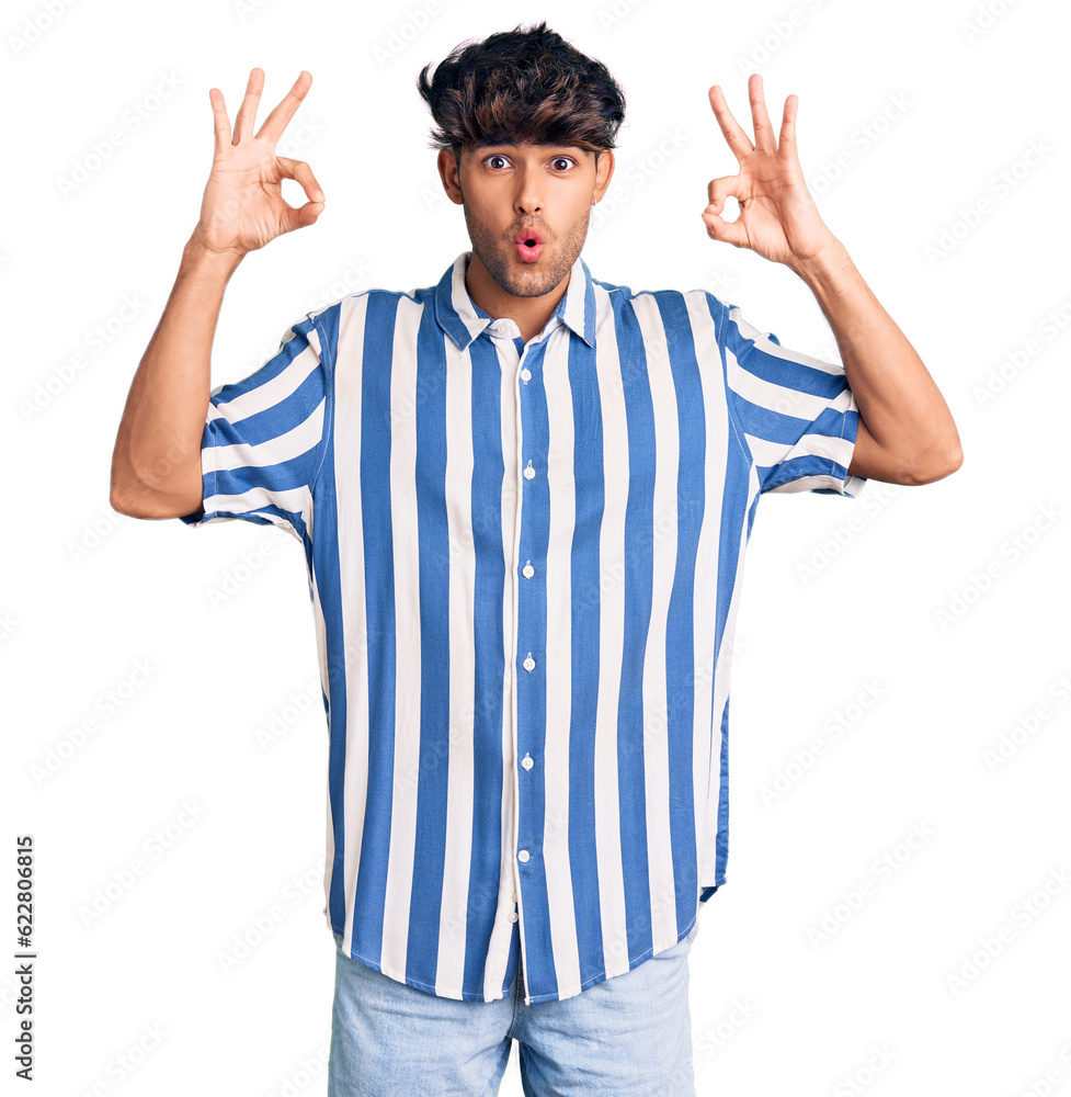 Young hispanic man wearing casual clothes looking surprised and shocked doing ok approval symbol with fingers. crazy expression Young hispanic man wearing casual clothes looking surprised and shocked doing ok approval symbol with fingers. crazy expression