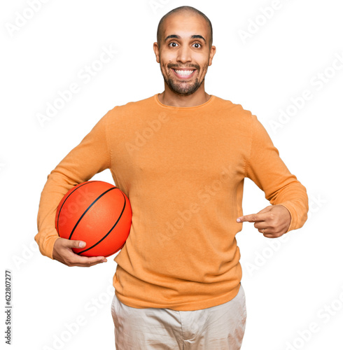 Hispanic adult man holding basketball ball smiling happy pointing with hand and finger