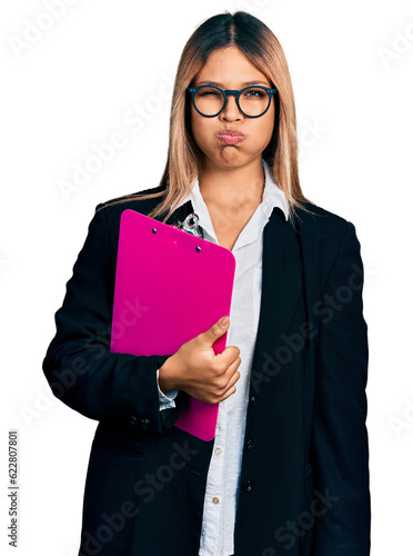 Young hispanic business woman holding clipboard with blank space puffing cheeks with funny face. mouth inflated with air, crazy expression.