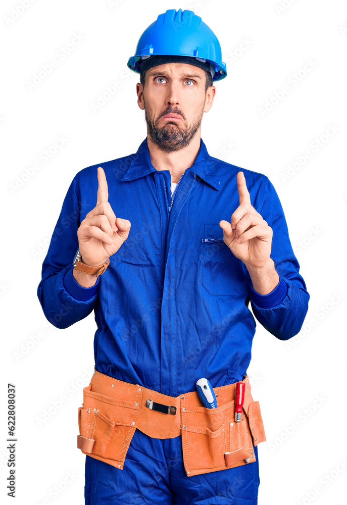 Young handsome man wearing worker uniform and hardhat pointing down looking sad and upset, indicating direction with fingers, unhappy and depressed.