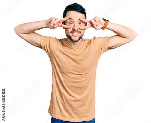 Hispanic man with beard wearing casual t shirt doing peace symbol with fingers over face, smiling cheerful showing victory