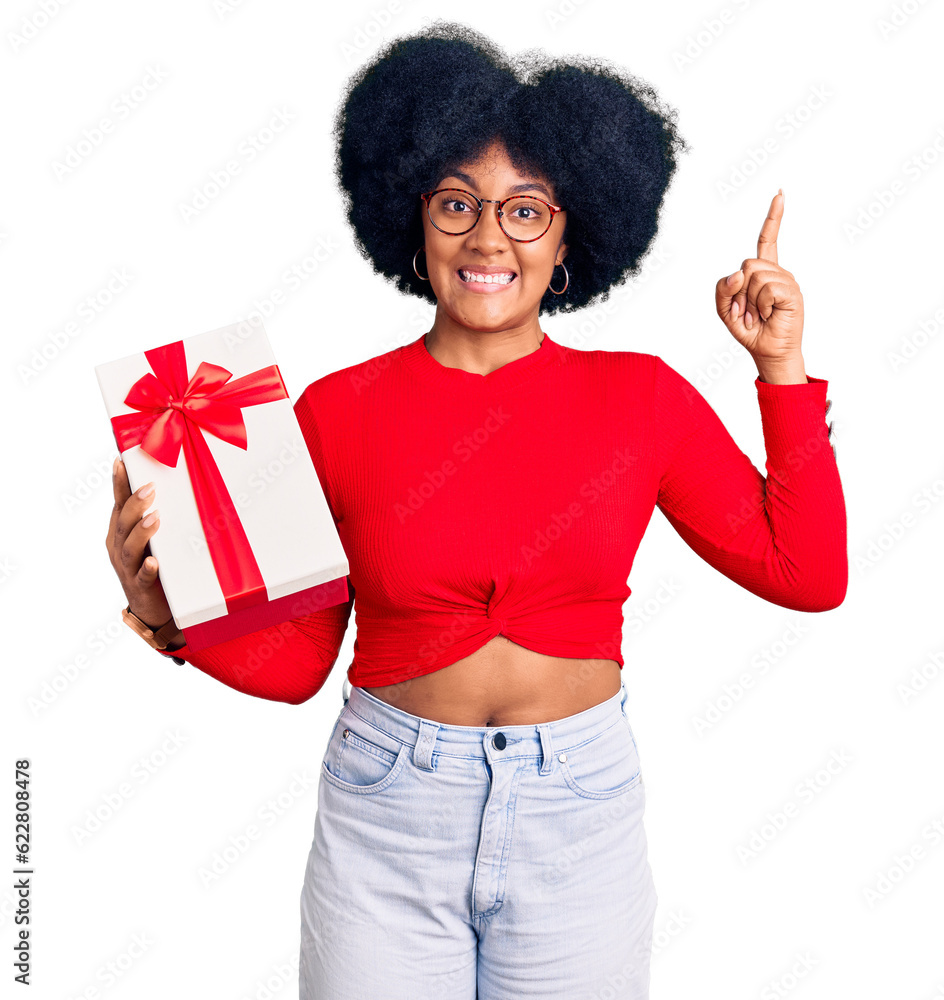 Young african american girl holding gift smiling amazed and surprised and pointing up with fingers and raised arms. Young african american girl holding gift smiling amazed and surprised and pointing up with fingers and raised arms.