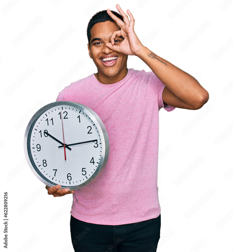 Young handsome hispanic man holding big clock smiling happy doing ok sign with hand on eye looking through fingers Young handsome hispanic man holding big clock smiling happy doing ok sign with hand on eye looking through fingers