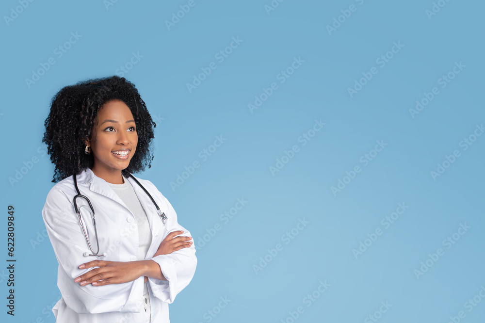 Happy young black woman doctor therapist in white coat with crossed arms on chest looks at free space Happy young black woman doctor therapist in white coat with crossed arms on chest looks at free space