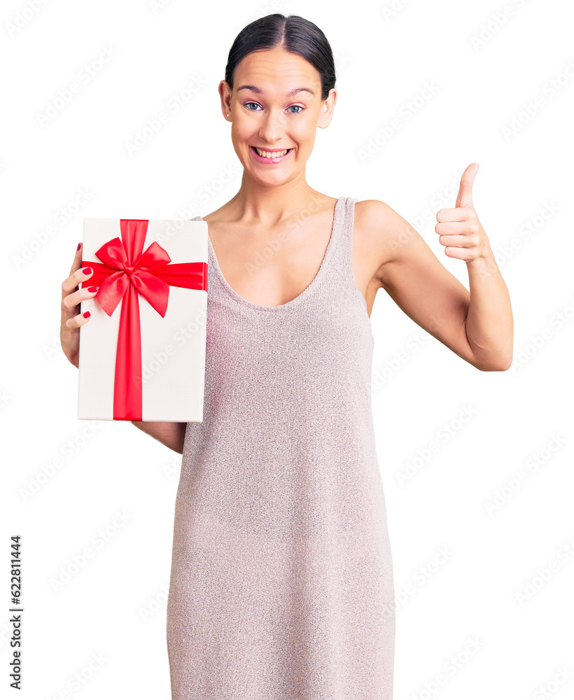 Beautiful brunette young woman holding gift smiling happy and positive, thumb up doing excellent and approval sign Beautiful brunette young woman holding gift smiling happy and positive, thumb up doing excellent and approval sign