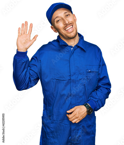 Bald man with beard wearing builder jumpsuit uniform waiving saying hello happy and smiling, friendly welcome gesture