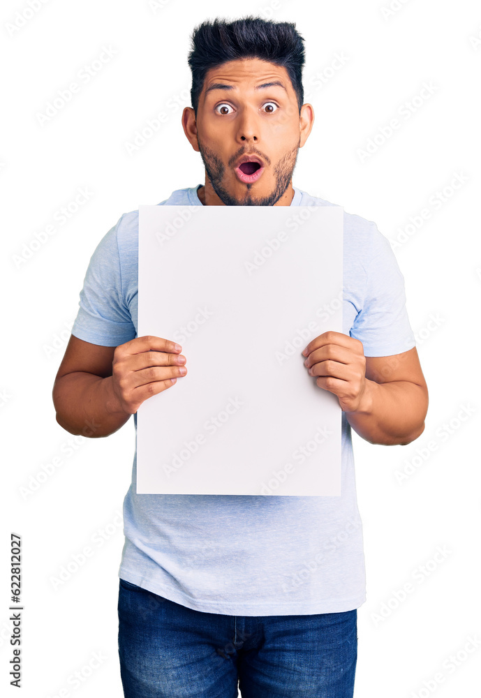 Handsome latin american young man holding cardboard banner with blank space scared and amazed with open mouth for surprise, disbelief face Handsome latin american young man holding cardboard banner with blank space scared and amazed with open mouth for surprise, disbelief face