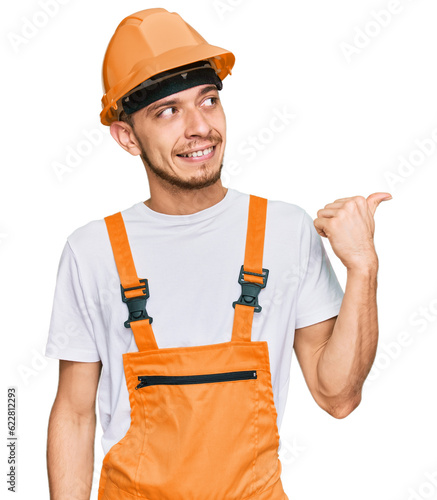 Hispanic young man wearing handyman uniform and safety hardhat smiling with happy face looking and pointing to the side with thumb up.