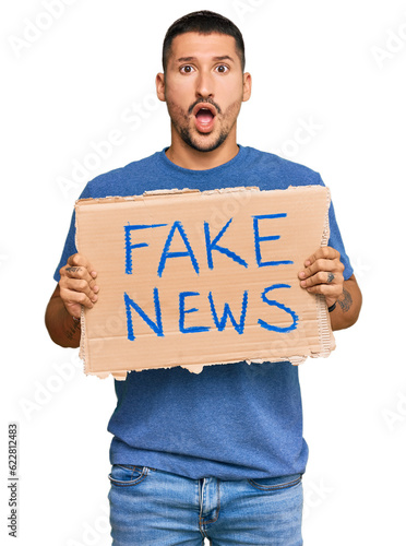 Handsome man with tattoos holding fake news banner scared and amazed with open mouth for surprise, disbelief face © Krakenimages.com