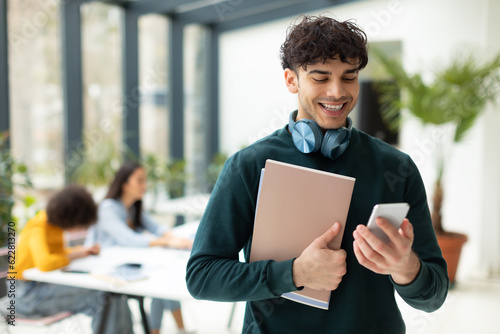 Cheerful european male student using smartphone, holding workbooks, standing in classroom with groupmates on background