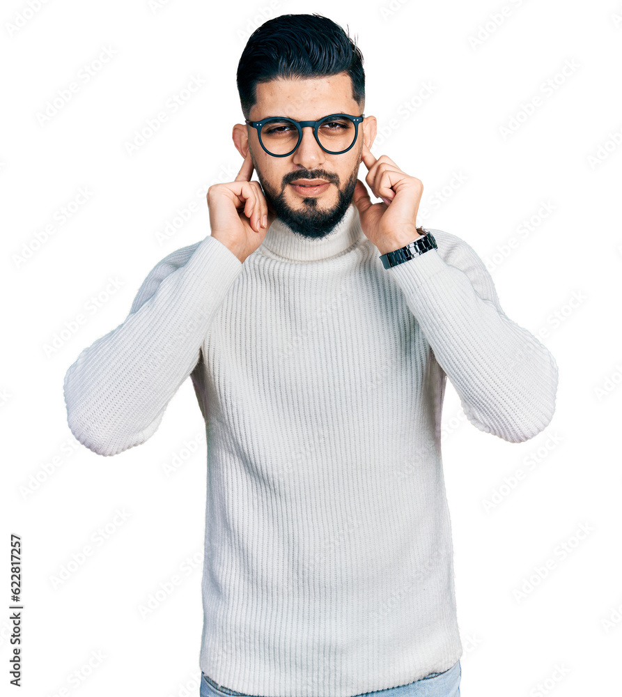 Fototapeta premium Young arab man with beard wearing elegant turtleneck sweater and glasses covering ears with fingers with annoyed expression for the noise of loud music. deaf concept.
