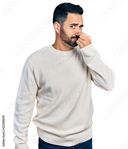 Young hispanic man with beard wearing casual white sweater smelling something stinky and disgusting, intolerable smell, holding breath with fingers on nose. bad smell photo