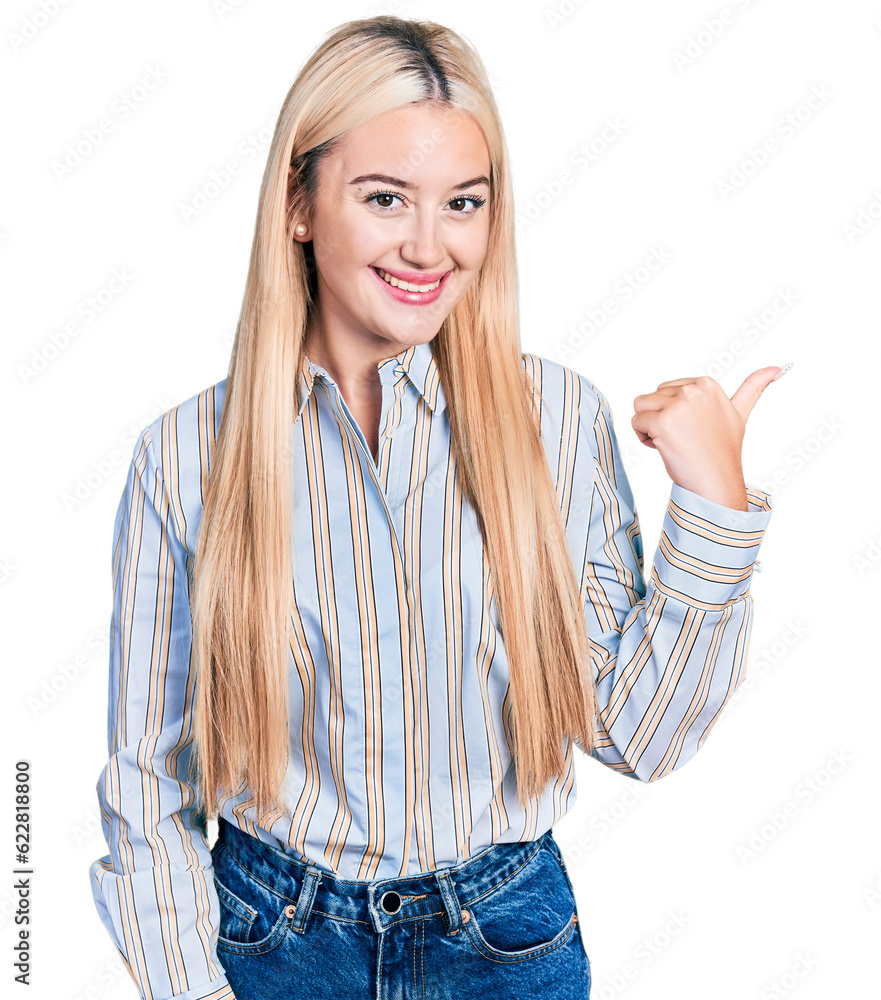 Beautiful blonde woman wearing casual striped shirt smiling with happy face looking and pointing to the side with thumb up. Beautiful blonde woman wearing casual striped shirt smiling with happy face looking and pointing to the side with thumb up.