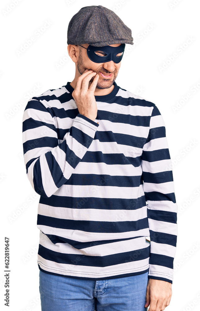 Young handsome man wearing burglar mask touching mouth with hand with painful expression because of toothache or dental illness on teeth. dentist Young handsome man wearing burglar mask touching mouth with hand with painful expression because of toothache or dental illness on teeth. dentist