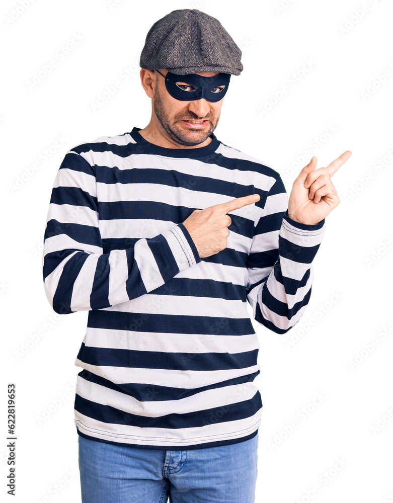 Young handsome man wearing burglar mask pointing aside worried and nervous with both hands, concerned and surprised expression Young handsome man wearing burglar mask pointing aside worried and nervous with both hands, concerned and surprised expression