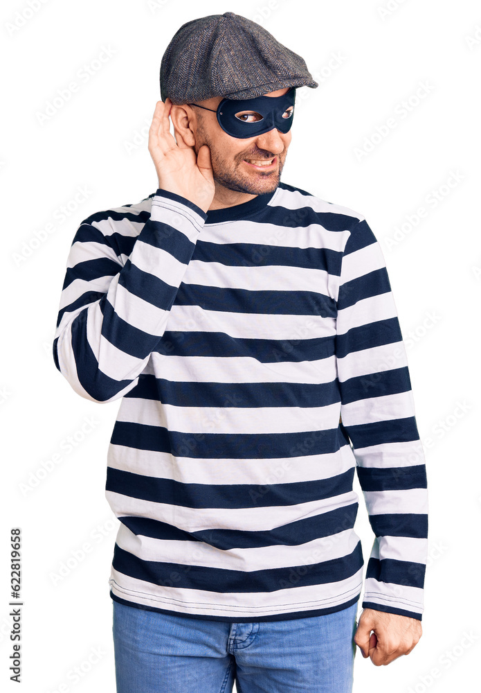 Young handsome man wearing burglar mask smiling with hand over ear listening an hearing to rumor or gossip. deafness concept. Young handsome man wearing burglar mask smiling with hand over ear listening an hearing to rumor or gossip. deafness concept.