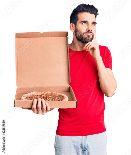 Young handsome man with beard holding delivery cardoboard with italian pizza serious face thinking about question with hand on chin, thoughtful about confusing idea photo