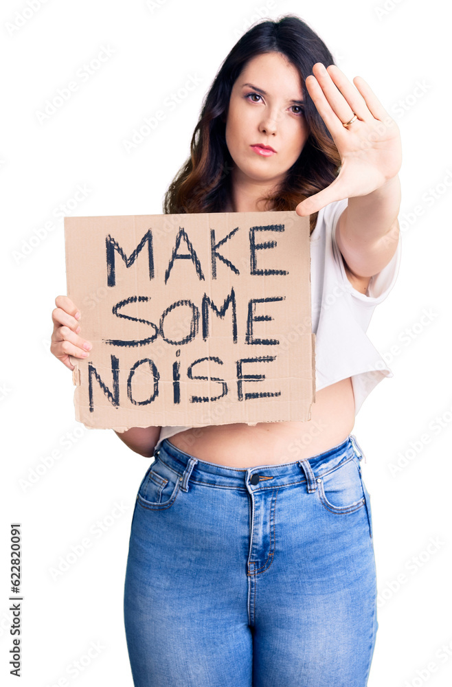 Beautiful young brunette woman holding make some noise banner with open hand doing stop sign with serious and confident expression, defense gesture Beautiful young brunette woman holding make some noise banner with open hand doing stop sign with serious and confident expression, defense gesture