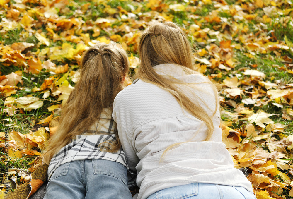Fototapeta premium The family has fun in the park. The girl and mother lie on a blanket with their backs to the camera and look at the autumn leaves.