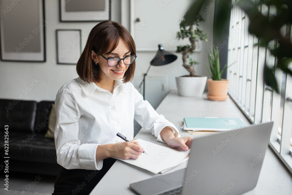 Smiling businesswoman using laptop and taking notes at workplace in modern office, lady watching business webinar Smiling businesswoman using laptop and taking notes at workplace in modern office, lady watching business webinar